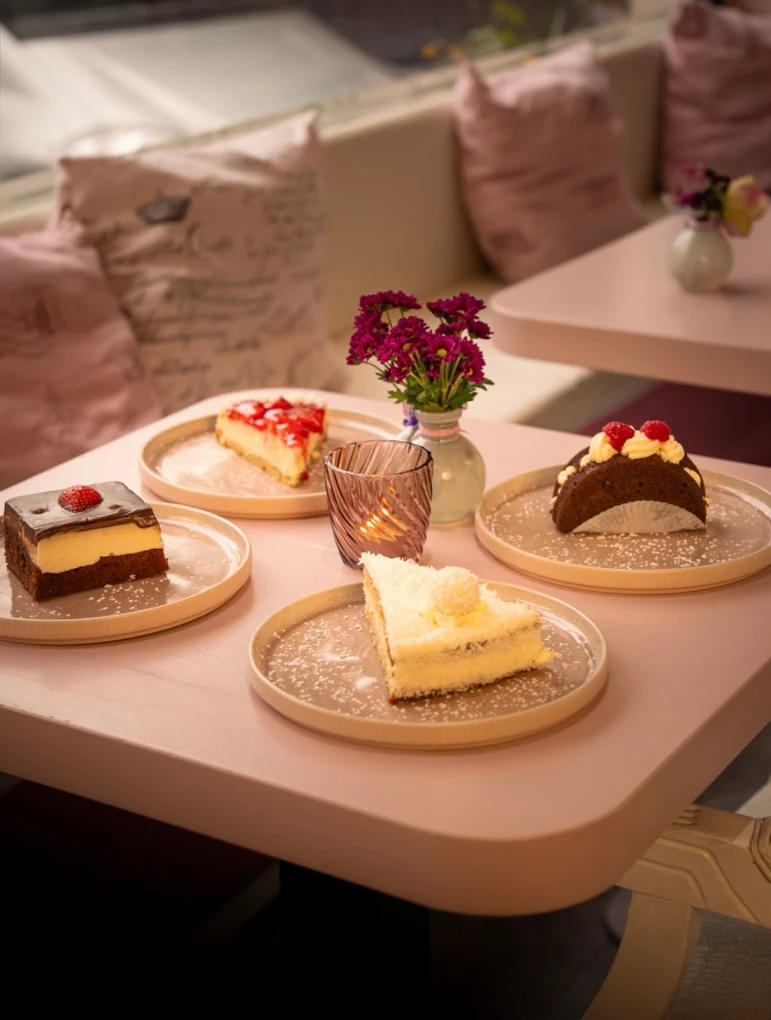 Vier Kuchenstücke auf Keramikteller mit Blumen und Kerze auf rosa Tisch